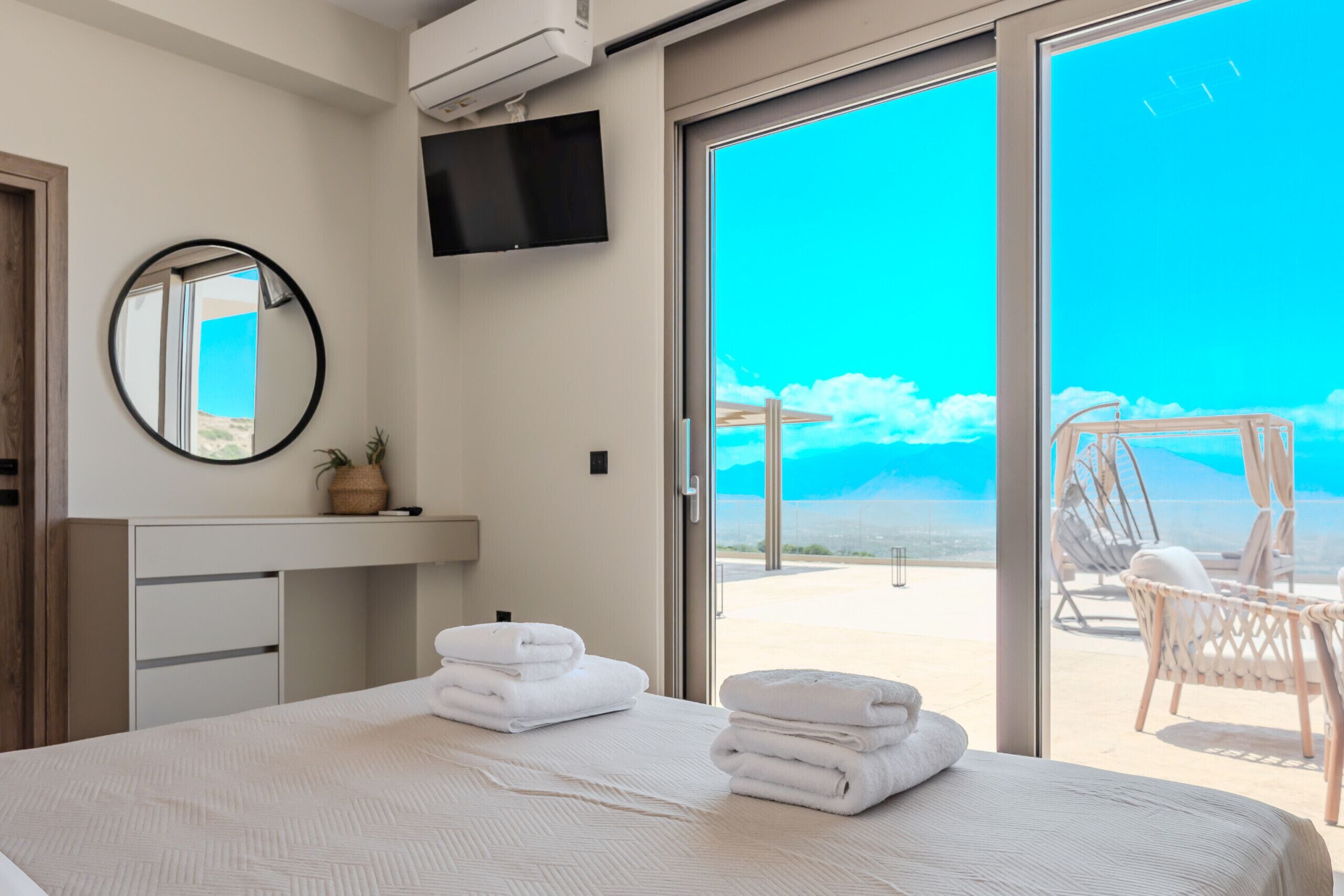 Une chambre à coucher comprenant un lit, un miroir rond, un téléviseur à écran plat et donnant sur un balcon équipé de chaises.