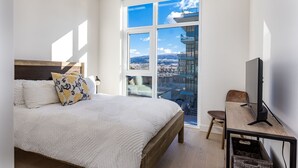 Ein Schlafzimmer mit einem großen Bett, einem Fernseher, einem Schreibtisch und Blick auf eine Stadtlandschaft durch ein großes Fenster.