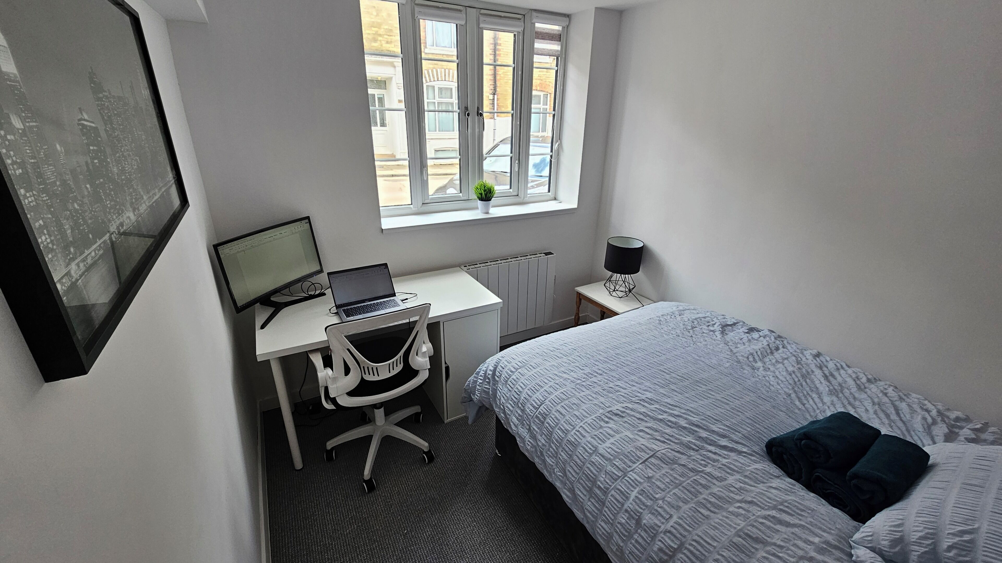Une chambre à coucher avec un lit, un bureau avec un ordinateur portable, une chaise, une fenêtre et un radiateur.