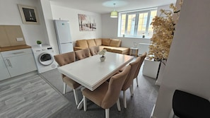 Ett modernt kök med en inbyggd tvättmaskin, en matplats med ett vitt bord och bruna stolar samt en liten sittgrupp i ett hörn med en soffa.