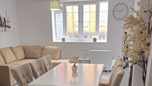 A modern living room with a beige sofa, a white dining table with chairs, a large window with curtains, and a clock on the wall. - Modern | Central | Park Free | Northants (West Northamptonshire)