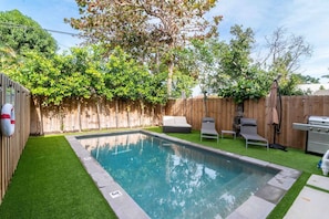Un patio trasero con piscina, césped artificial, cerca de madera y mobiliario exterior.