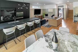 Una cocina moderna con una isla central, taburetes de bar, una mesa de comedor con un florero y una sala de estar con sofá y televisor.