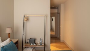 Ein Hotelzimmer mit einem Bett, einem Schreibtisch mit Lampe, einem Tablett mit Flasche und Gläsern sowie einem Korridor, der in ein weiteres Zimmer führt.