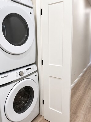 Uma área de lavanderia com duas máquinas de lavar Samsung brancas, uma porta fechada e piso de madeira.