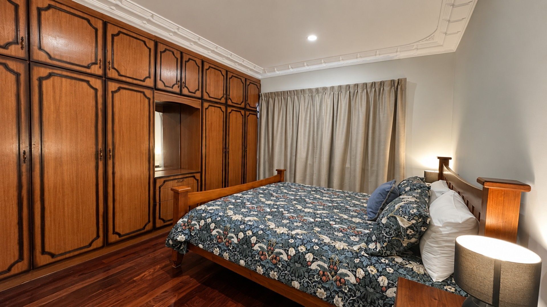 A bedroom with a wooden wardrobe, a bed with floral bedding, a nightstand, and a lamp.