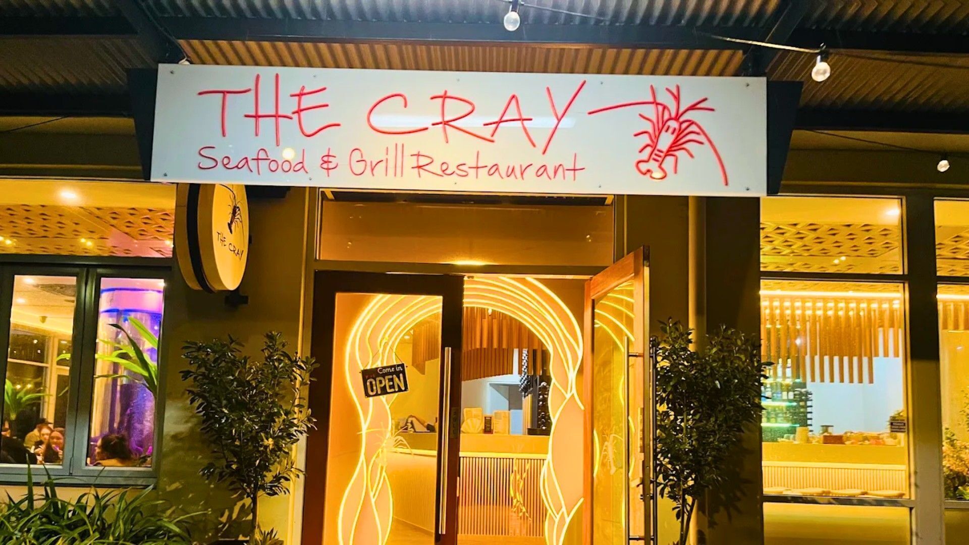 The exterior of The Cray Seafood & Grill Restaurant with a neon sign and a glass door entrance.