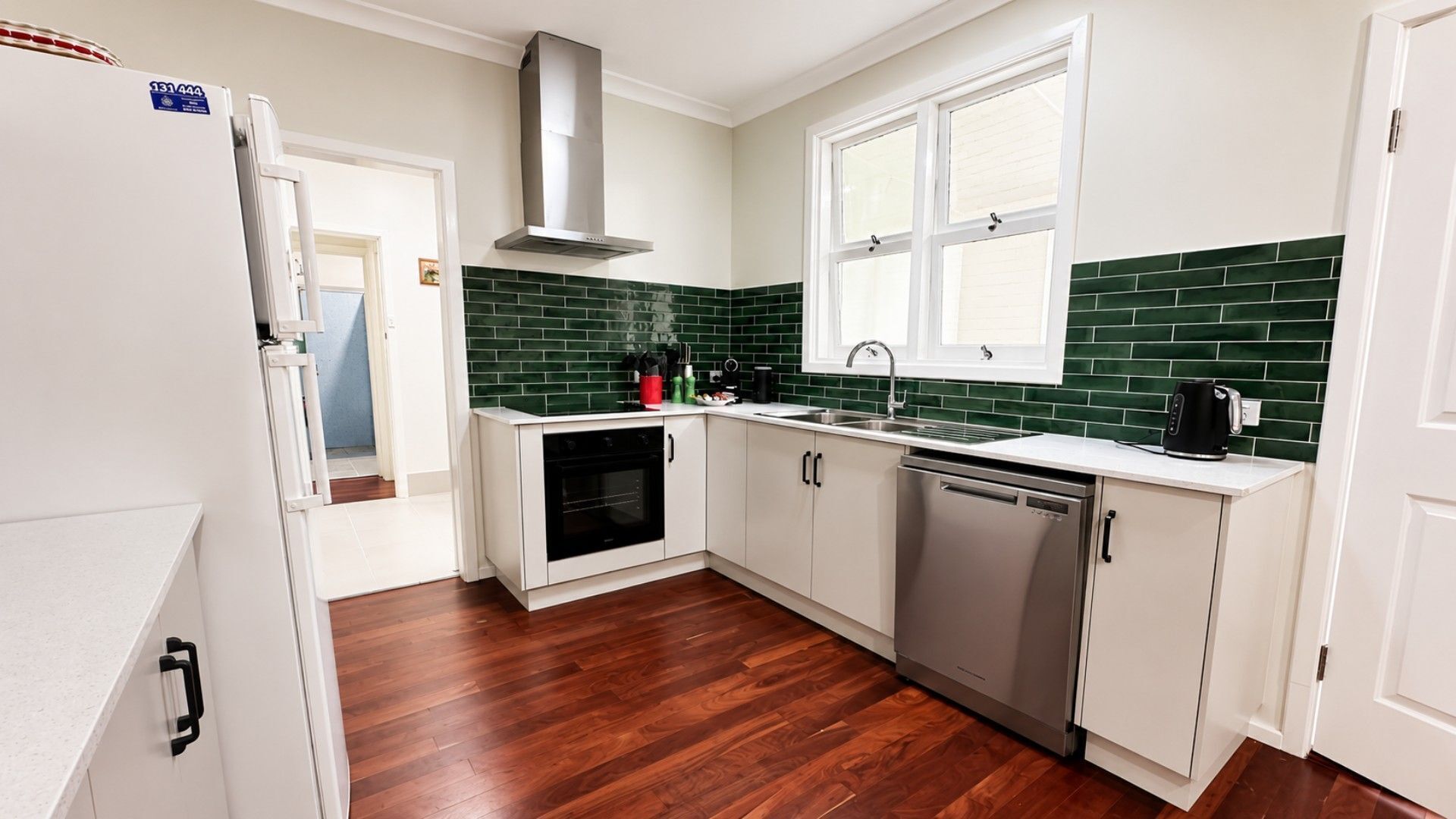 A kitchen with white cabinets, a stainless steel dishwasher, a built-in oven, a window with blinds, and a green-tiled backsplash.