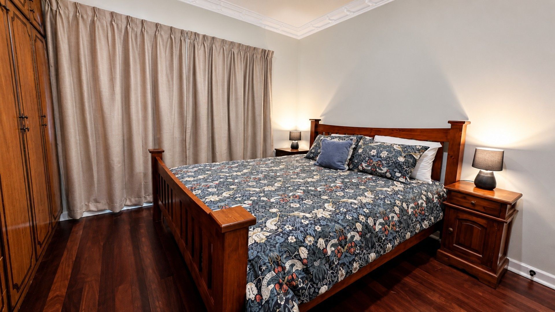 A bedroom with a wooden bed, a nightstand with a lamp, a wardrobe, and a large window with curtains.