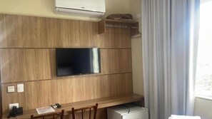 Ett rum med en väggmonterad platt-TV, väggpaneler i trä, ett skrivbord med en stol, ett litet kylskåp och ett fönster med gardiner.