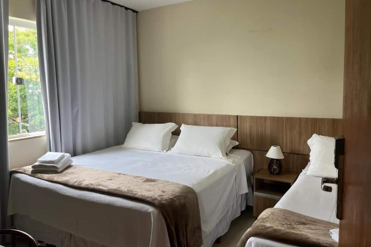 Ett hotellrum med en säng, ett nattduksbord, en lampa och ett fönster med gardiner.