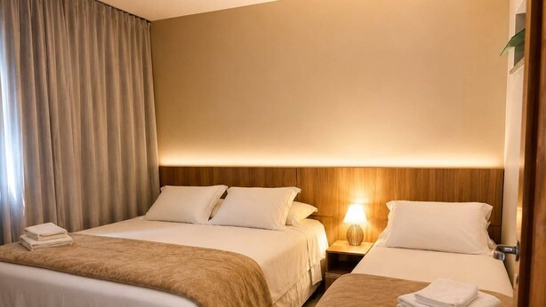 A hotel room with two beds, a wooden headboard, a bedside table with a lamp, and a window with curtains.