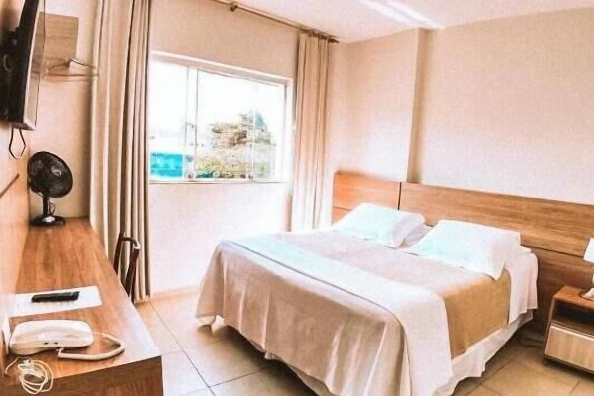 Ett hotellrum med en säng, ett nattduksbord, en lampa och ett fönster med gardiner.
