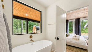 Een moderne badkamer met een vrijstaand bad, een houten plafond en een groot raam met uitzicht op een bos.