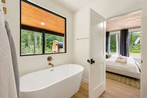 Ein modernes Badezimmer mit freistehender Badewanne, Holzboden und einem großen Fenster mit Blick auf einen Wald.