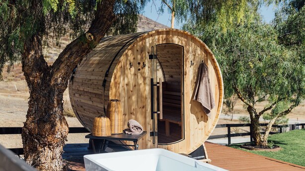 Une structure en forme de tonneau, faite de bois, avec une porte, une petite table sur laquelle se trouve un panier, et un banc blanc à l’extérieur.
