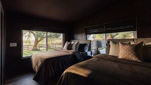 Une chambre avec deux lits, une table de chevet, une lampe et une vue sur un arbre à l’extérieur.