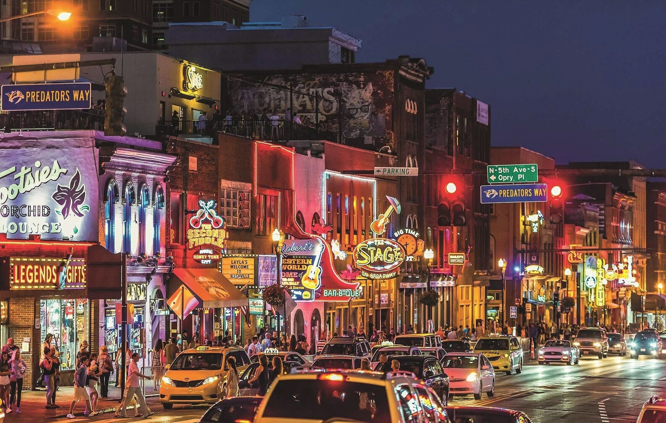 Une rue animée d’une ville, de nuit, avec diverses devantures de magasins et enseignes illuminées, notamment « Predators Way » et « Legends Gifts ».