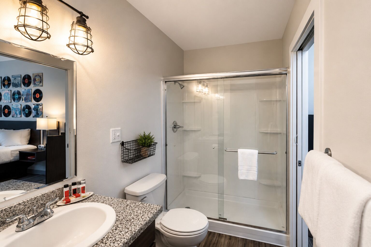 A modern bathroom with a glass shower enclosure, a white toilet, a granite countertop sink, and a mirror with wall-mounted lights.