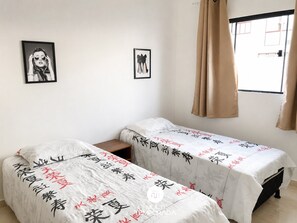 Quarto com duas camas, cada uma com colcha branca com texto vermelho e preto. Há duas fotos emolduradas na parede, uma janela com cortinas e uma pequena mesa de cabeceira de madeira entre as camas.