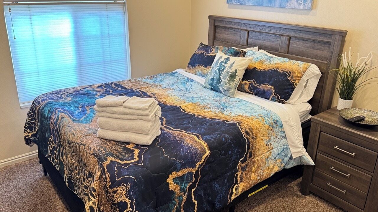 A bed with a patterned bedspread, a bedside table with a plant, and a window with blinds.