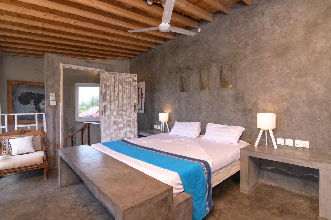 A bedroom with a bed, bedside lamps, a wooden bench, a ceiling fan, and a view through an open door.