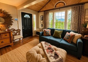 A cozy living room with a dark blue sofa, a wooden chair, a patterned rug, and a large window with curtains. - Guthrie Ridge-Charming Getaway, Gorgeous Views! (Waynesville)