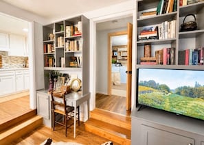 A modern kitchen with white cabinetry, a dining area with a wooden table and chairs, and a bookshelf filled with books and decorative items. - Guthrie Ridge-Charming Getaway, Gorgeous Views! (Waynesville)