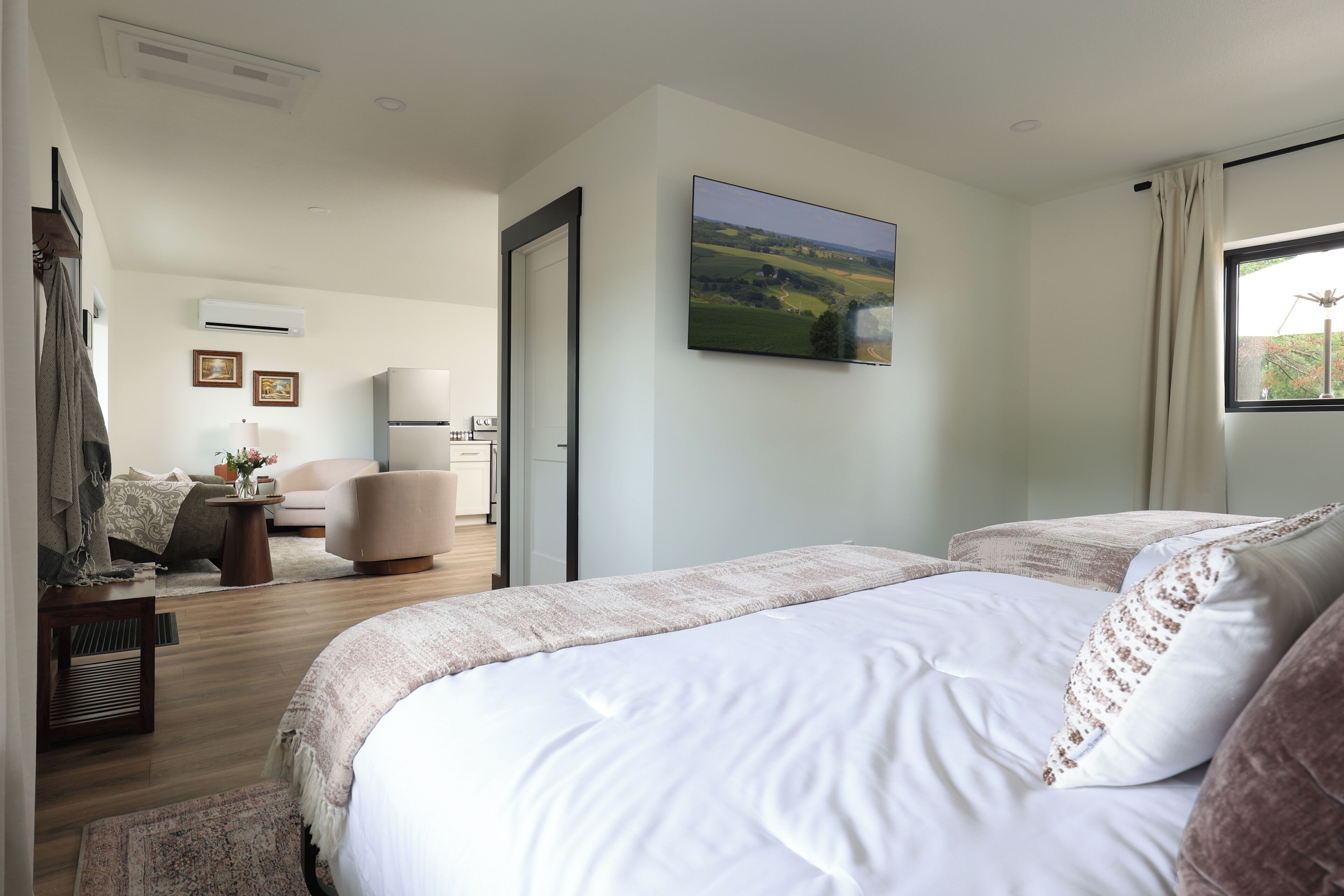 A hotel room with a large bed, a flat-screen TV mounted on the wall, and a view of a green landscape through the window.