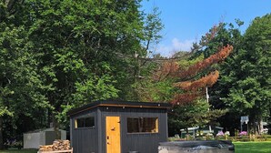 A small wooden cabin with a door, windows, and a hot tub in a grassy area with trees. - Steps from the Lake- Sauna + Hot Tub. Unit #2 (Jones)