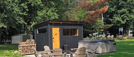 Una pequeña cabaña de madera con puerta, ventanas y un jacuzzi en un área verde con árboles.