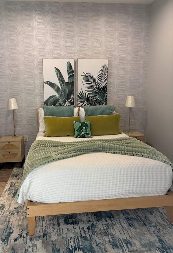 A neatly made bed with a green and white bedspread, two pillows, and a matching throw blanket. There are two bedside lamps on wooden nightstands, a patterned wallpaper, and a large plant on the wall.