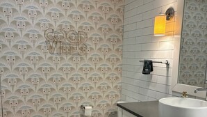 Kylpyhuoneessa on kuviollinen tapetti, wc-istuin, peilillä varustettu pesuallas ja puinen jakkara.