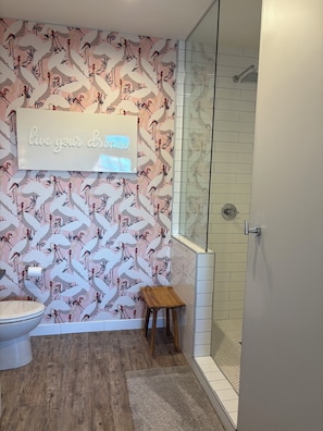 A bathroom with a patterned wall, a toilet, a small wooden stool, and a glass shower enclosure.
