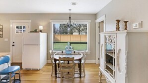 A kitchen with a dining table, chairs, a refrigerator, and a window with a view of a fence and trees. - Thompson Cottage - Sleeps 4 (Sedro-Woolley)
