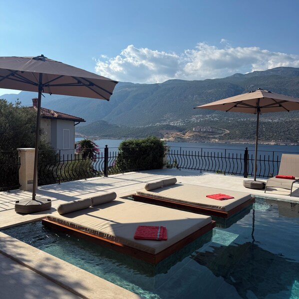 An infinity pool with lounge chairs and umbrellas, a view of a mountainous lake, and a fenced patio area.