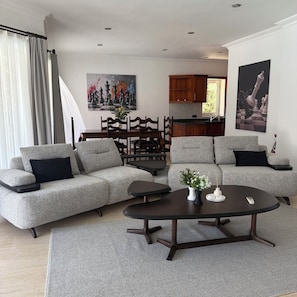 A modern living room with a grey sofa, a coffee table, and a dining area with wooden chairs and a kitchen in the background.