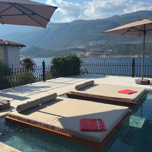 A poolside area with lounge chairs and umbrellas, overlooking a body of water and mountains.