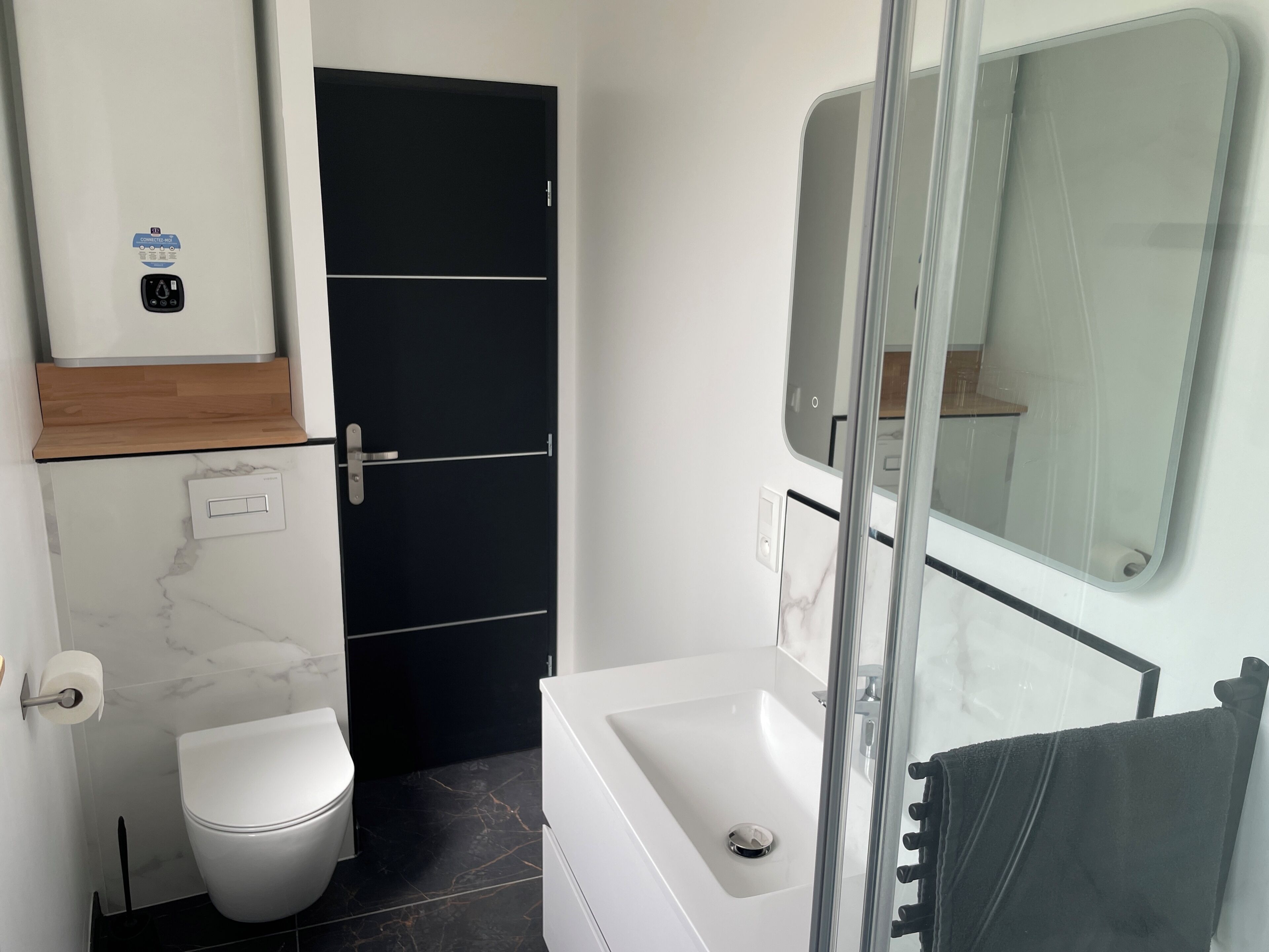 Une salle de bain comprenant des toilettes, une baignoire, un espace de douche vitré, une porte noire et un lavabo blanc.