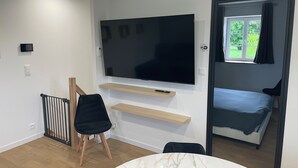 Et moderne værelse med et fladskærms-TV monteret på væggen, en seng ved et vindue, et lille bord og en stol.