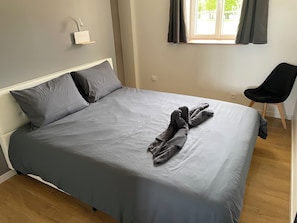A neatly made bed with grey bedding, a black chair, and a window with curtains.