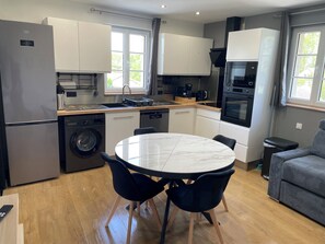 A modern kitchen with a built-in washing machine, a dining table with chairs, and a grey sofa.