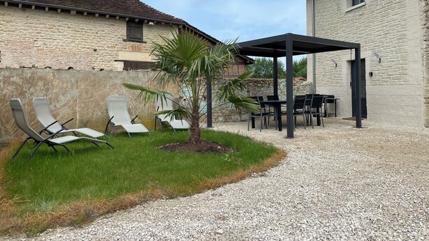 En terrasse med udendørs møbler, en lille palme og en grussti, der fører til en bygning med stenvæg.