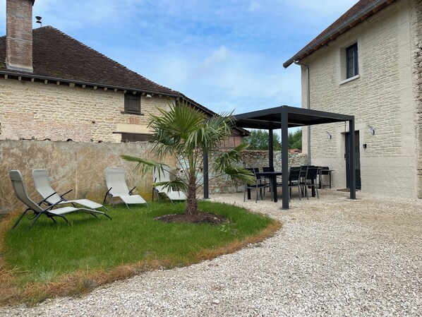A patio area with outdoor furniture, a small palm tree, and a gravel path leading to a building with a stone facade.