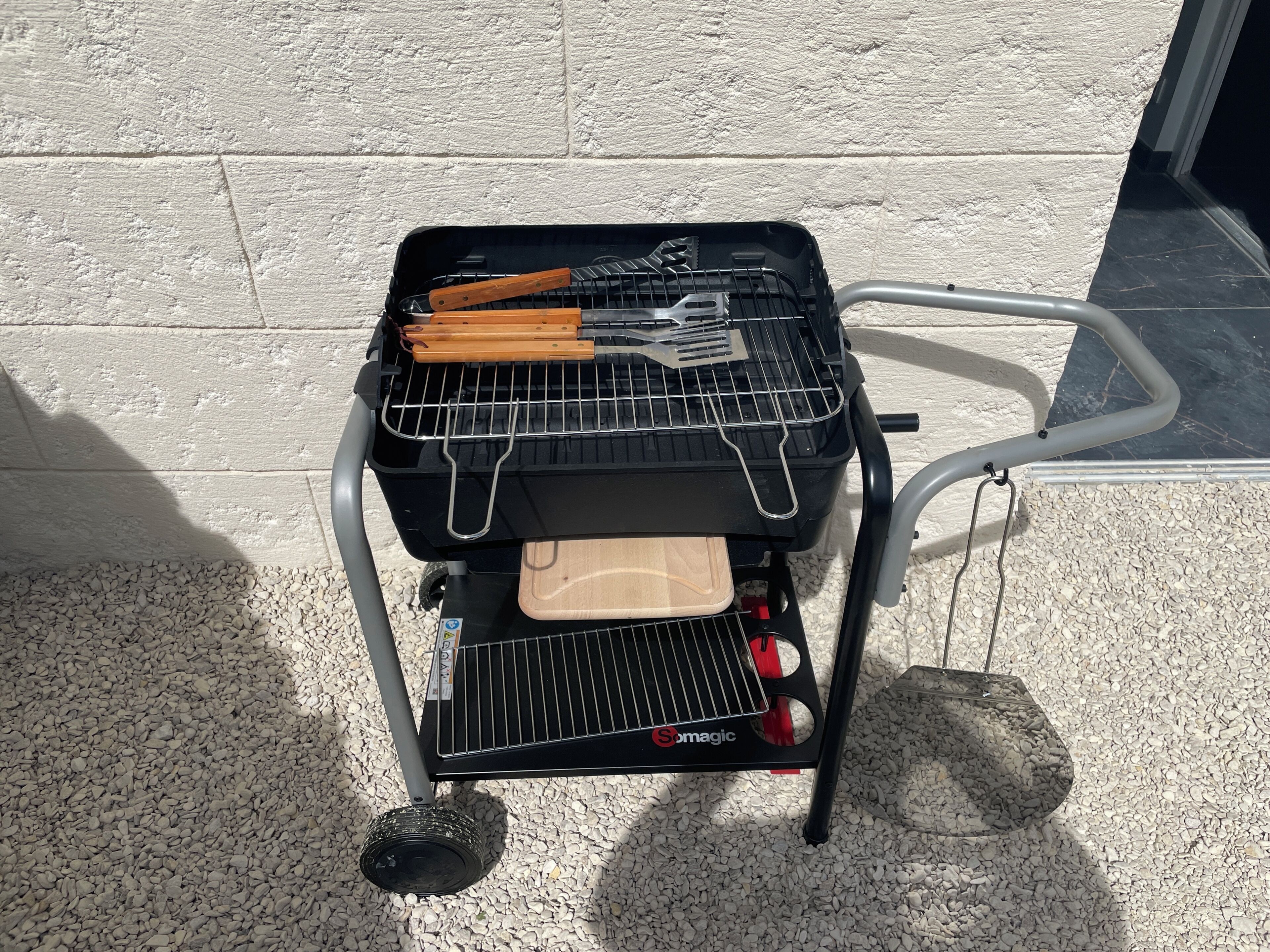 A portable grill with a lid, a spatula, tongs, and a tray, placed on a gravel surface against a white wall.