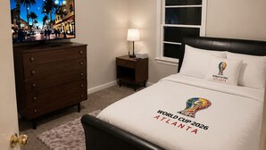 Une chambre à coucher comprenant un lit, une commode, une table de chevet, une lampe, un téléviseur et une fenêtre.