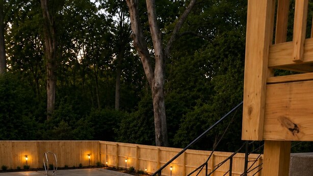 Un escalier en béton avec des rampes métalliques mène à une terrasse en bois, qui est entourée d’une clôture en bois et éclairée par des luminaires encastrés dans le sol.