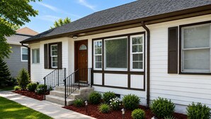 Une maison de plain-pied avec un porche à l’avant, une porte en bois et un petit jardin devant, agrémenté d’arbustes et d’un chemin.
