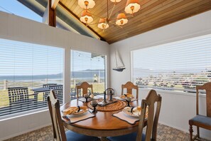 A dining area with a table set for four, large windows with blinds, a sailboat decoration, and a view of a coastal landscape. - Little Blue - BEACH FRONT on Samish Island (Bow)