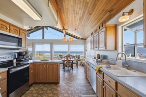 A modern kitchen with wooden cabinets, a central island, a large window, and a view of the ocean. - Little Blue - BEACH FRONT on Samish Island (Bow)